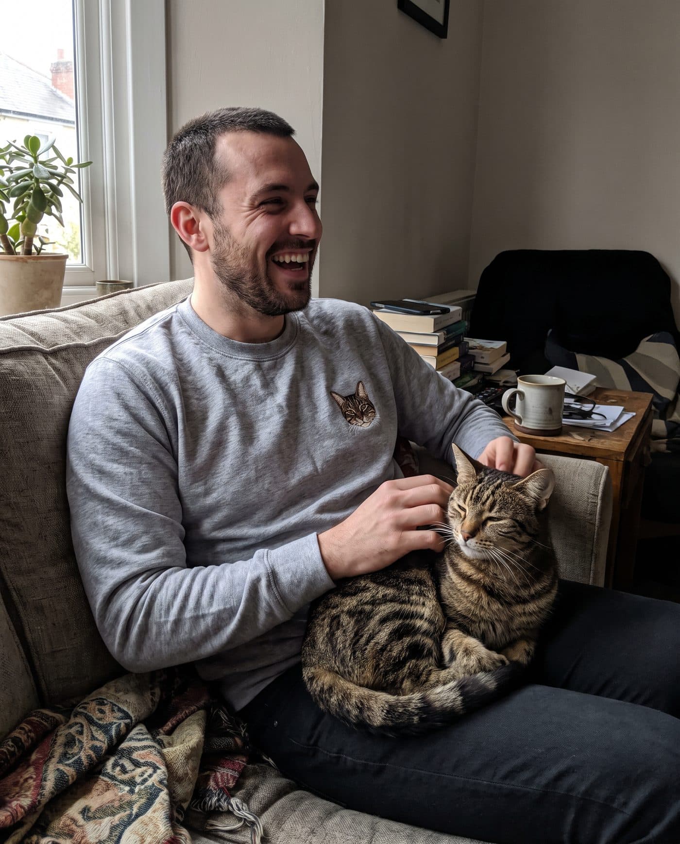 A man on his sofa in a grey marl sweatshirt with his tabby cat's portrait embroidered on it, scratching the cat on his lap
